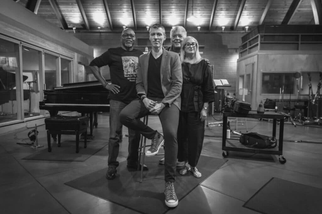 Black and white photo of the Legacy band: Lewis Nash, Nick Finzer, Rufus Reid, and Renee Rosnes. They stand in the recording studio looking directly at the camera.