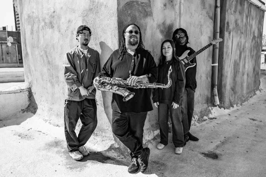 Black and white photo of Jongkuk Kim, Dayna Stephens, Kanoa Mendenhall, and Emmanuel Michael standing against a concrete backdrop.