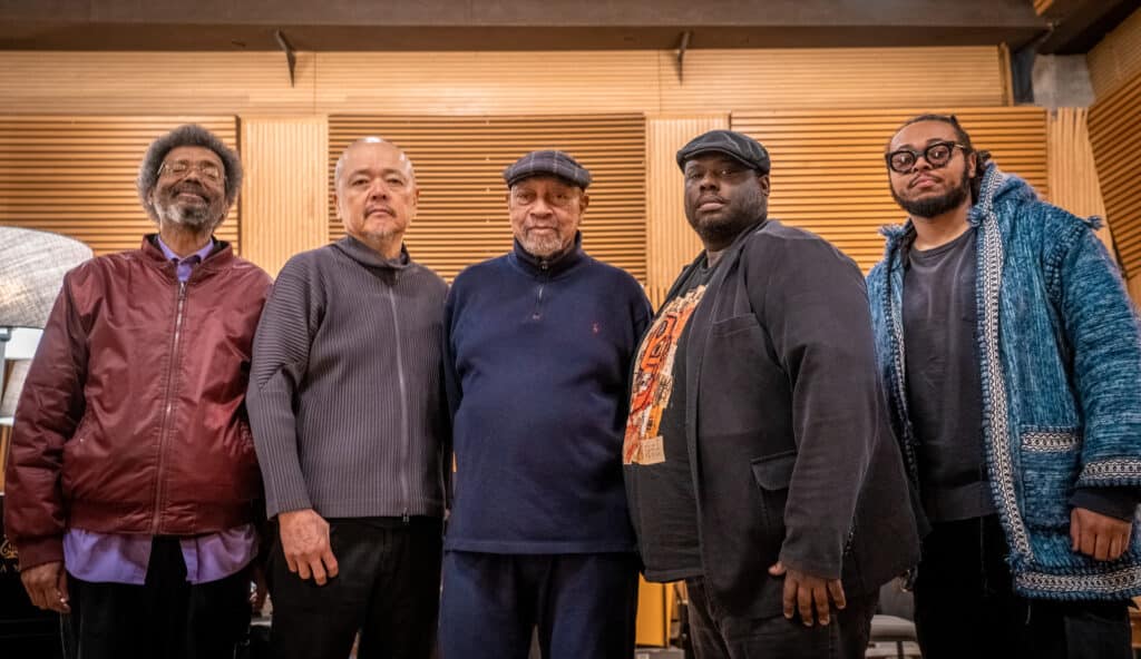 The full band stand in a row in the studio, arms around each other, looking straight into the camera.