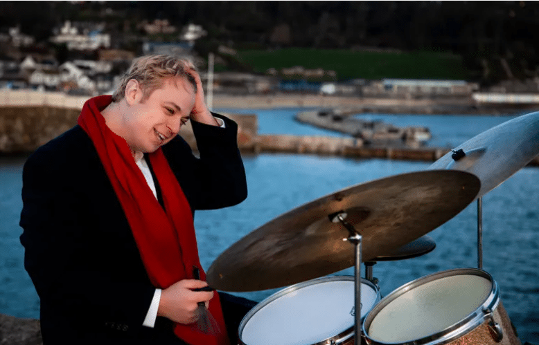 Douglas Marriner smiles and runs a hand through his hair, sitting at a drum kit outside with a harbour scene in the background.