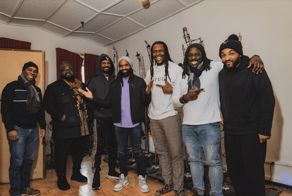 The whole band, 7 musicians, stand in a row, smiling in the studio. Obed Calvaire is in the middle holding his hands outstretched.