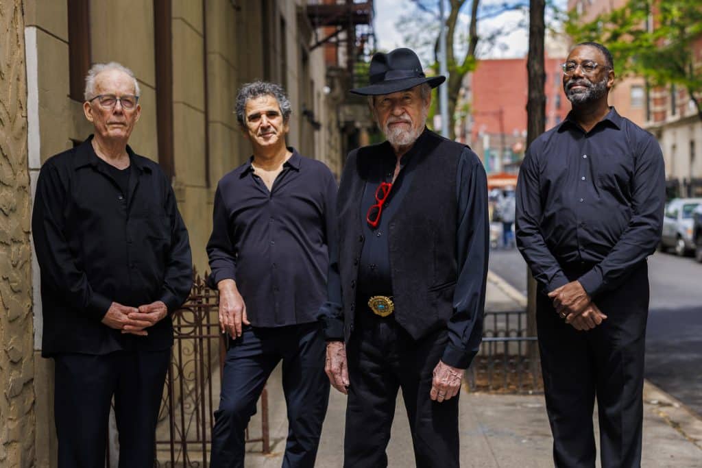 The four musicians stand in a row on a New York City street, wearing all black, looking into the camera and smiling.