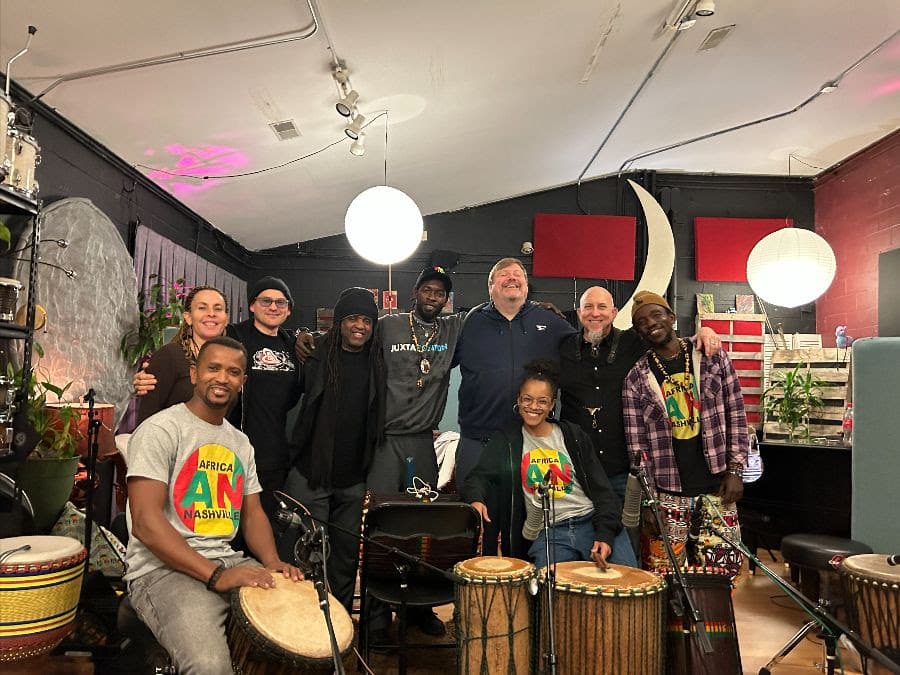 A group of nine musicians stand in a group, smiling with their arms around each other, surrounded by drums.