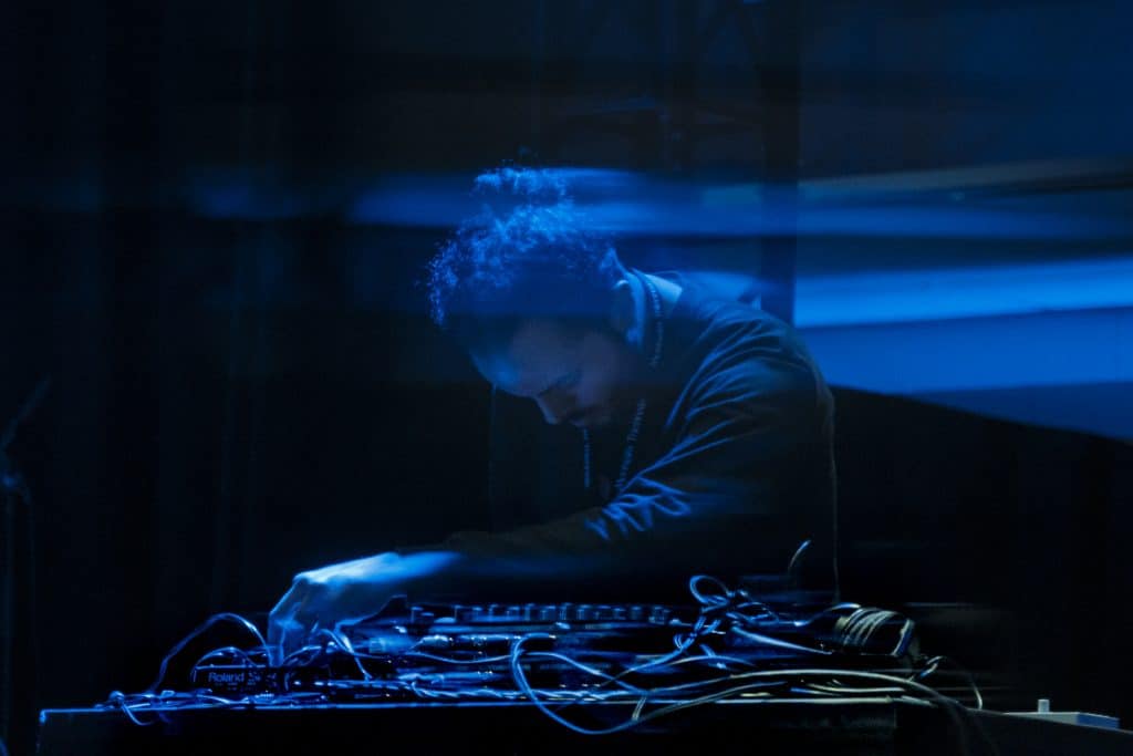 Muqat'a leans over a sound board, looking intently down during a concert. The background is black with blue lighting.