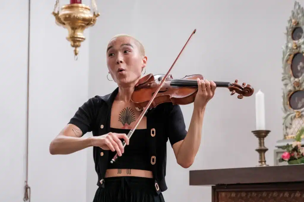 yuniya edi kwon plays the violin in a white-painted room with ornate decorations. Her face is animated, looking off to the side.