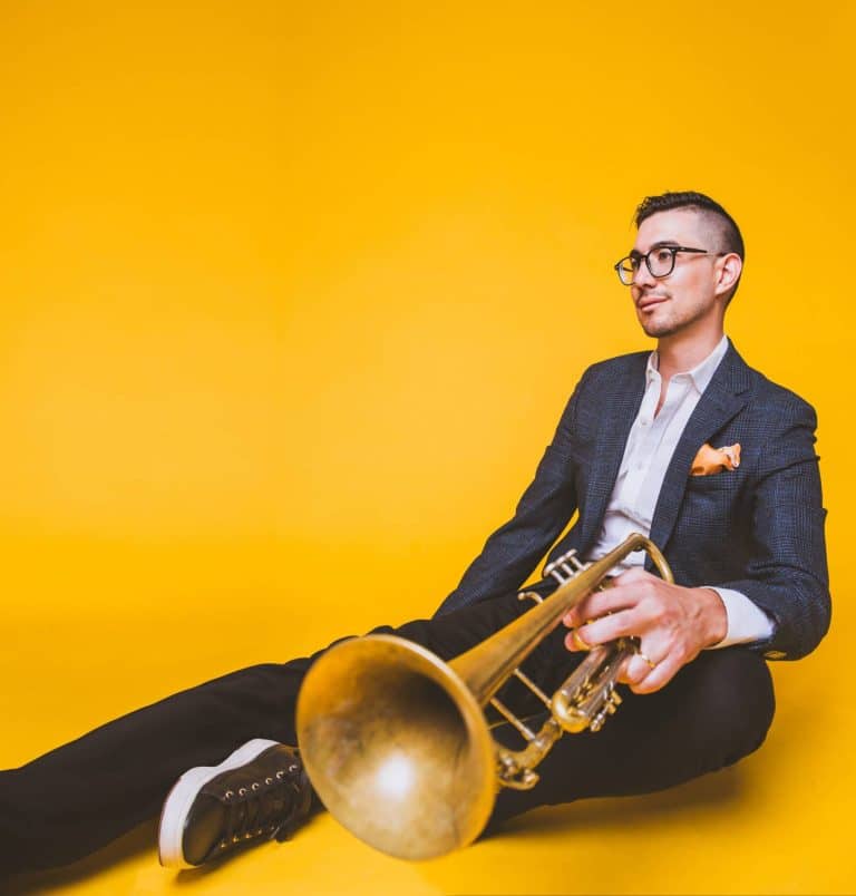 Danny Jonokuchi sits on the ground against a bright yellow background, looking off to the side and holding his trombone in the foreground.