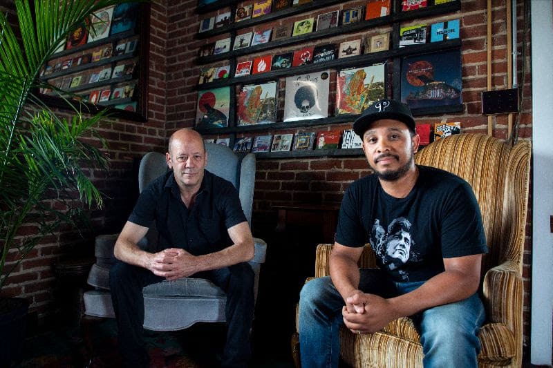 The two men sit in armchairs against an exposed brick wall displaying various CDs and records.