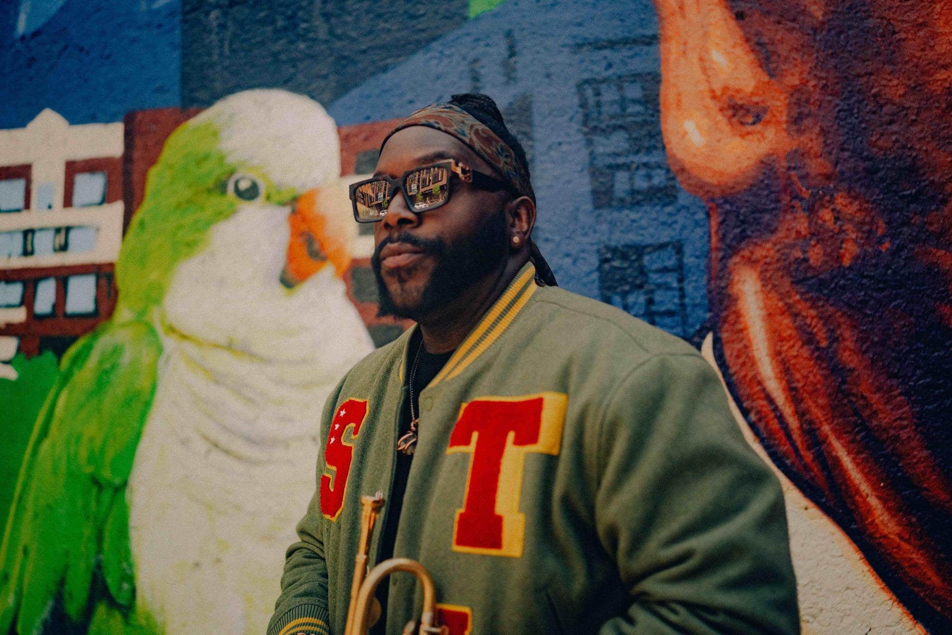 Marquis Hill stands against a brightly painted street wall featuring a green parrot. He wears shades and looks at the camera slightly side-on.