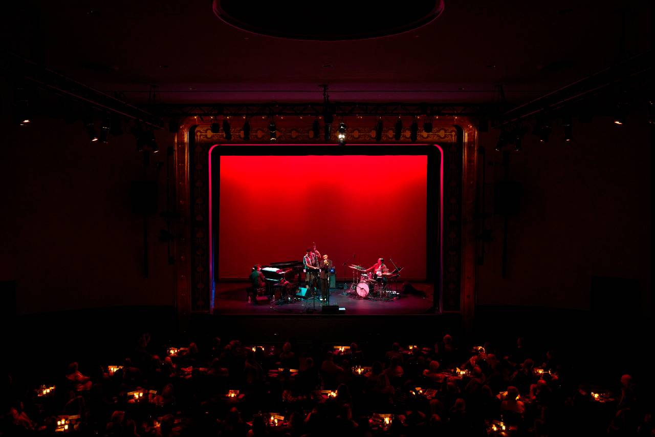 A shot of a stage from the back of a large room, with a red curtain at the back and a quartet playing. The stage is surrounded by cabaret tables.