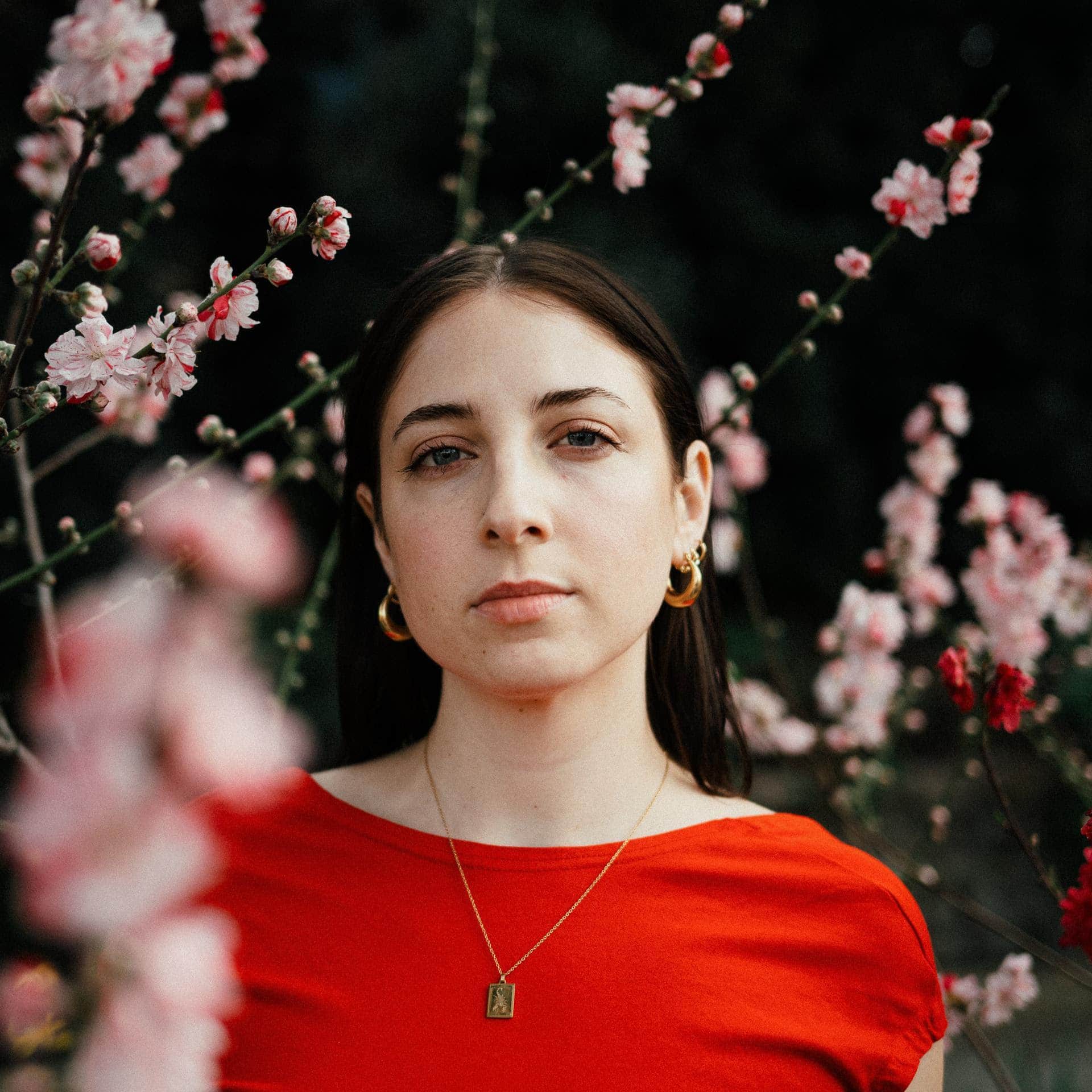 Alden Hellmuth stands among pink blossoms and looks directly into the camera, wearing a red dress.