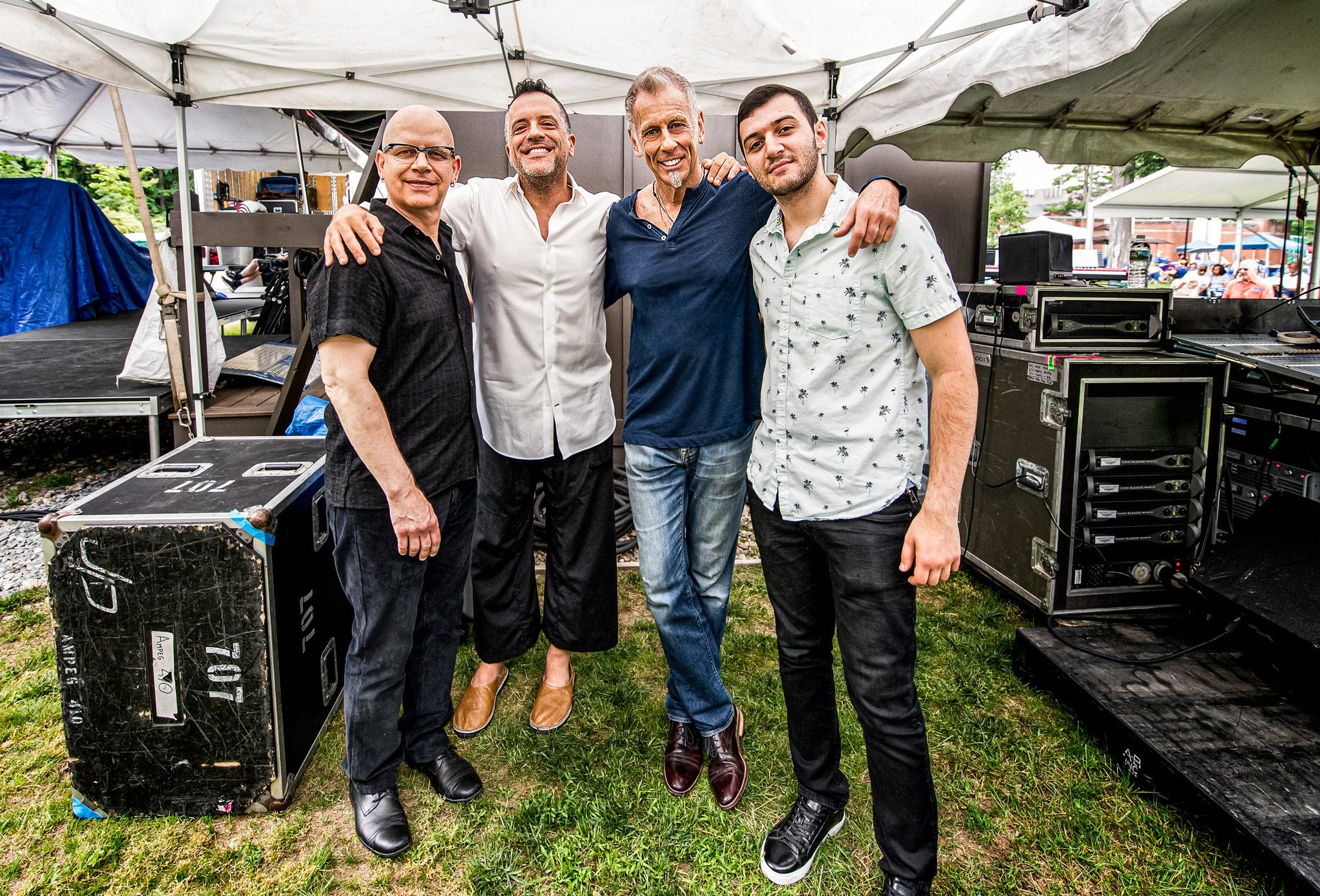 Four male musicians stand in a row with their arms around each other, smiling into the camera. 