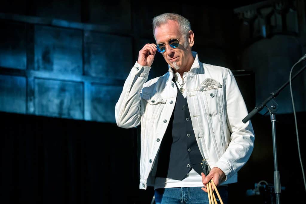 Joe Locke stands on a stage wearing sunglasses. He is wearing a white jacket and holding his sticks.